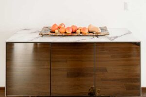 Komoda s lesklými dvířky v dřevěném dekoru a kamennou deskou s dekorací.