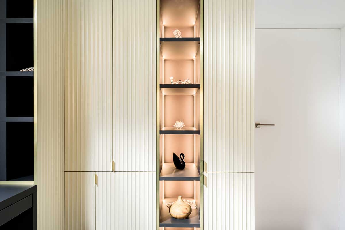 Vestavěná vitrína s vertikálním podsvícením a dekorativními policemi zapuštěnými do skříňové stěny.