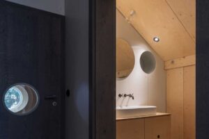 Vstup do koupelny s dřevěnými dveřmi kruhovým okénkem a světlým interiérem