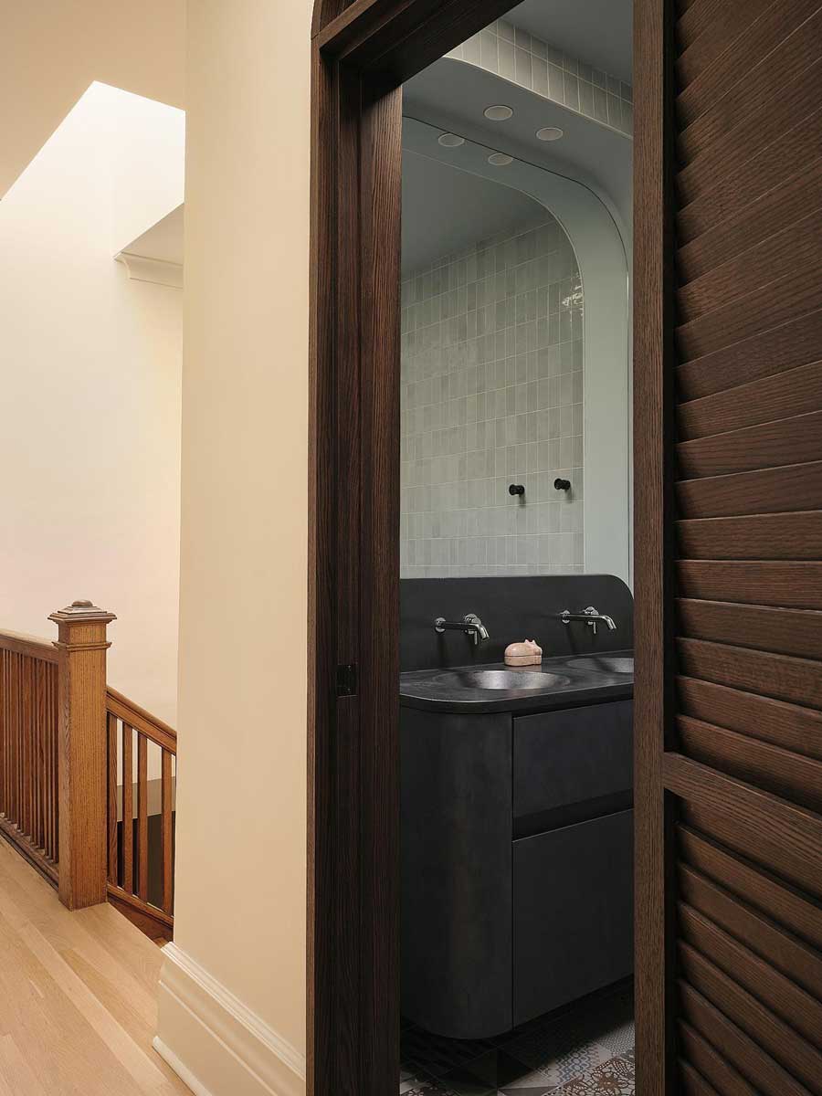 Vstup do koupelny s tmavým vestavěným umyvadlem, zaobleným průchodem a původním dřevěným schodištěm