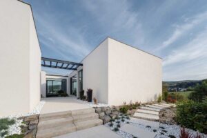 Vstupní cesta k modernímu domu s bílou fasádou, kamennými schody a upravenou zahradou.