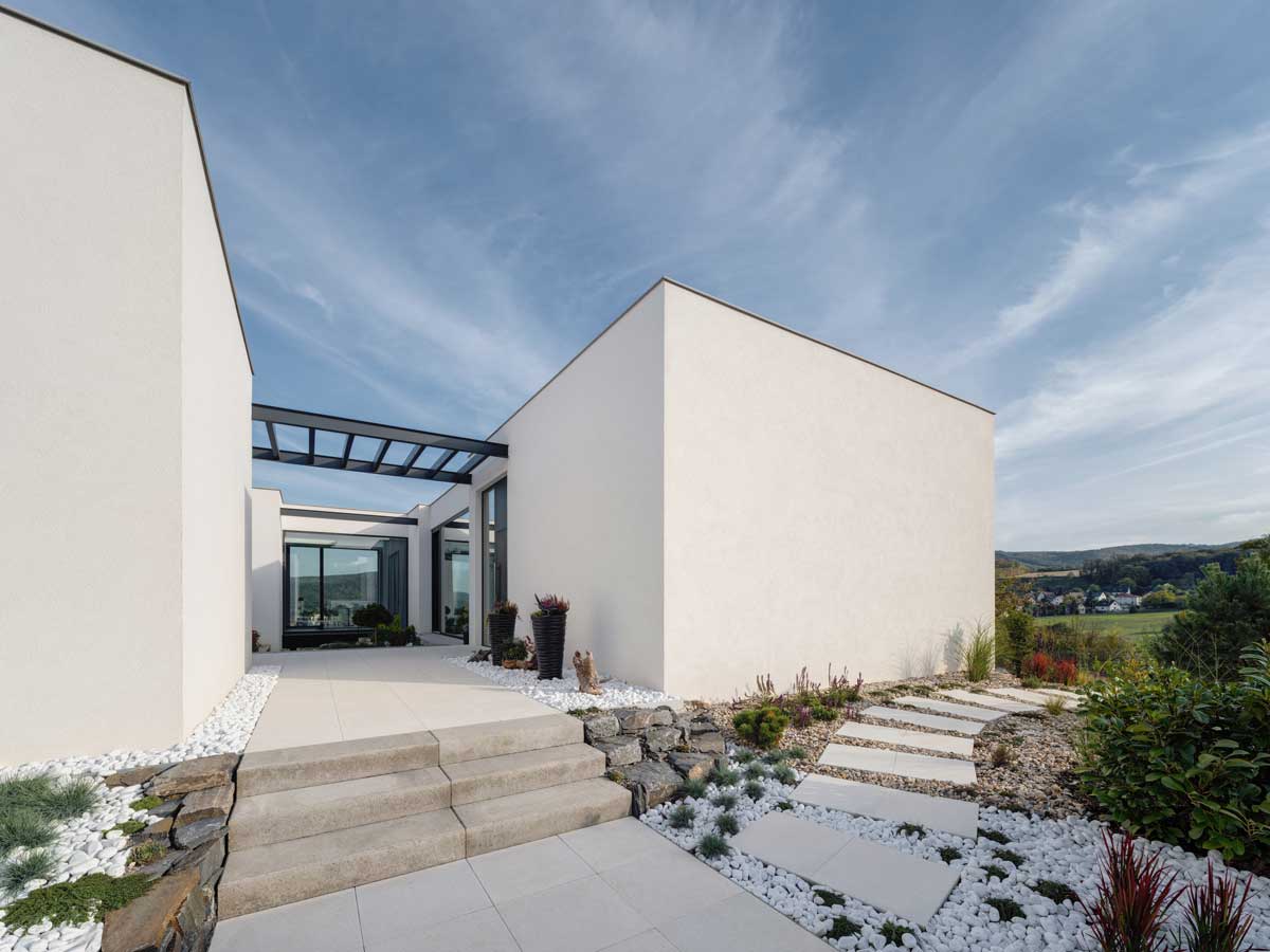 Vstupní cesta k modernímu domu s bílou fasádou, kamennými schody a upravenou zahradou.