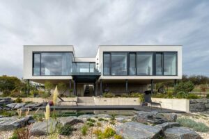 Moderní patrový rodinný dům s velkoformátovými okny, betonovým soklem a přírodním koupacím jezírkem v upravené zahradě.