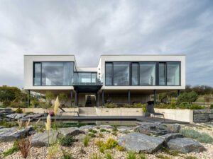 Moderní patrový rodinný dům s velkoformátovými okny, betonovým soklem a přírodním koupacím jezírkem v upravené zahradě.