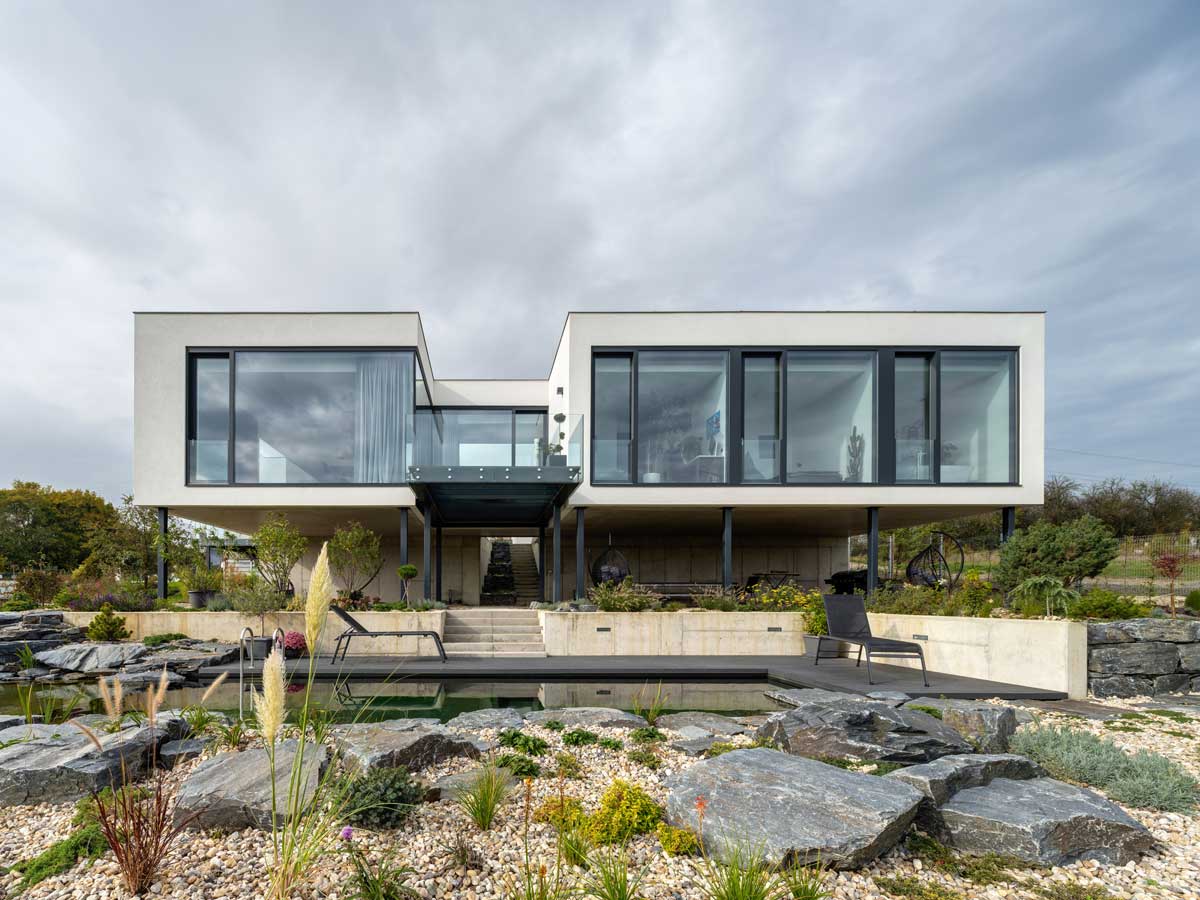 Moderní patrový rodinný dům s velkoformátovými okny, betonovým soklem a přírodním koupacím jezírkem v upravené zahradě.