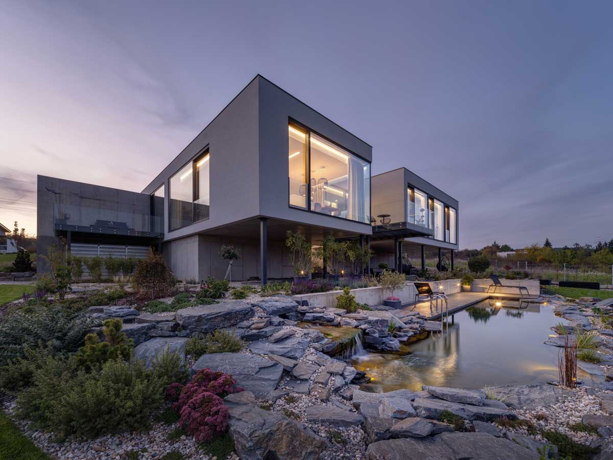 Moderní rodinný dům s prosklenou fasádou a zahradou při večerním nasvícení.