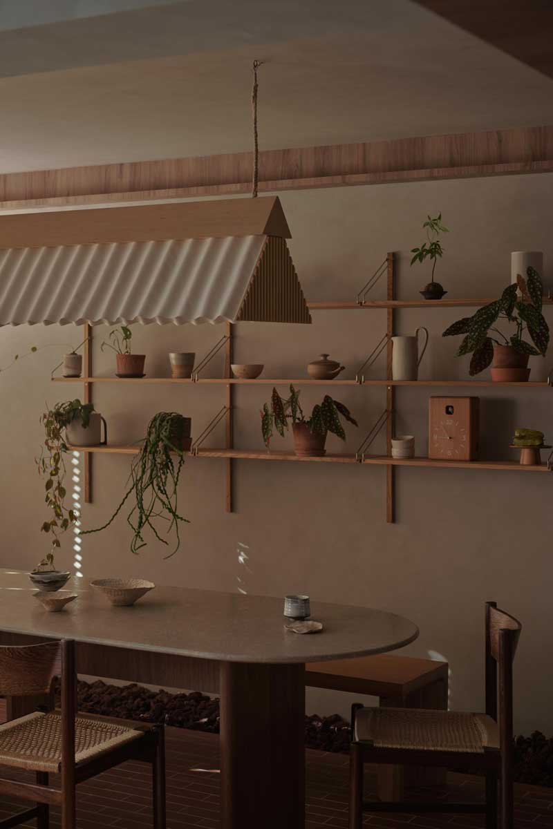 Oválný jídelní stůl z tmavého dřeva je obklopen jednoduchými židlemi a otevřenými policemi s rostlinami a keramikou.