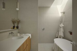 Minimalistická koupelna v Lilac House s vestavěnou vanou, světlými obklady a dřevěnou skříňkou pod umyvadlem.