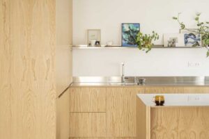 Minimalistická kuchyň s dřevěnými skříňkami na míru, nerezovou pracovní deskou a přiznaným betonovým stropem po rekonstrukci bytu