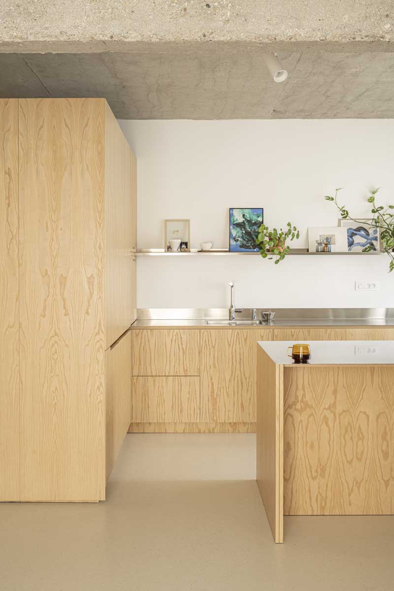 Minimalistická kuchyň s dřevěnými skříňkami na míru, nerezovou pracovní deskou a přiznaným betonovým stropem po rekonstrukci bytu