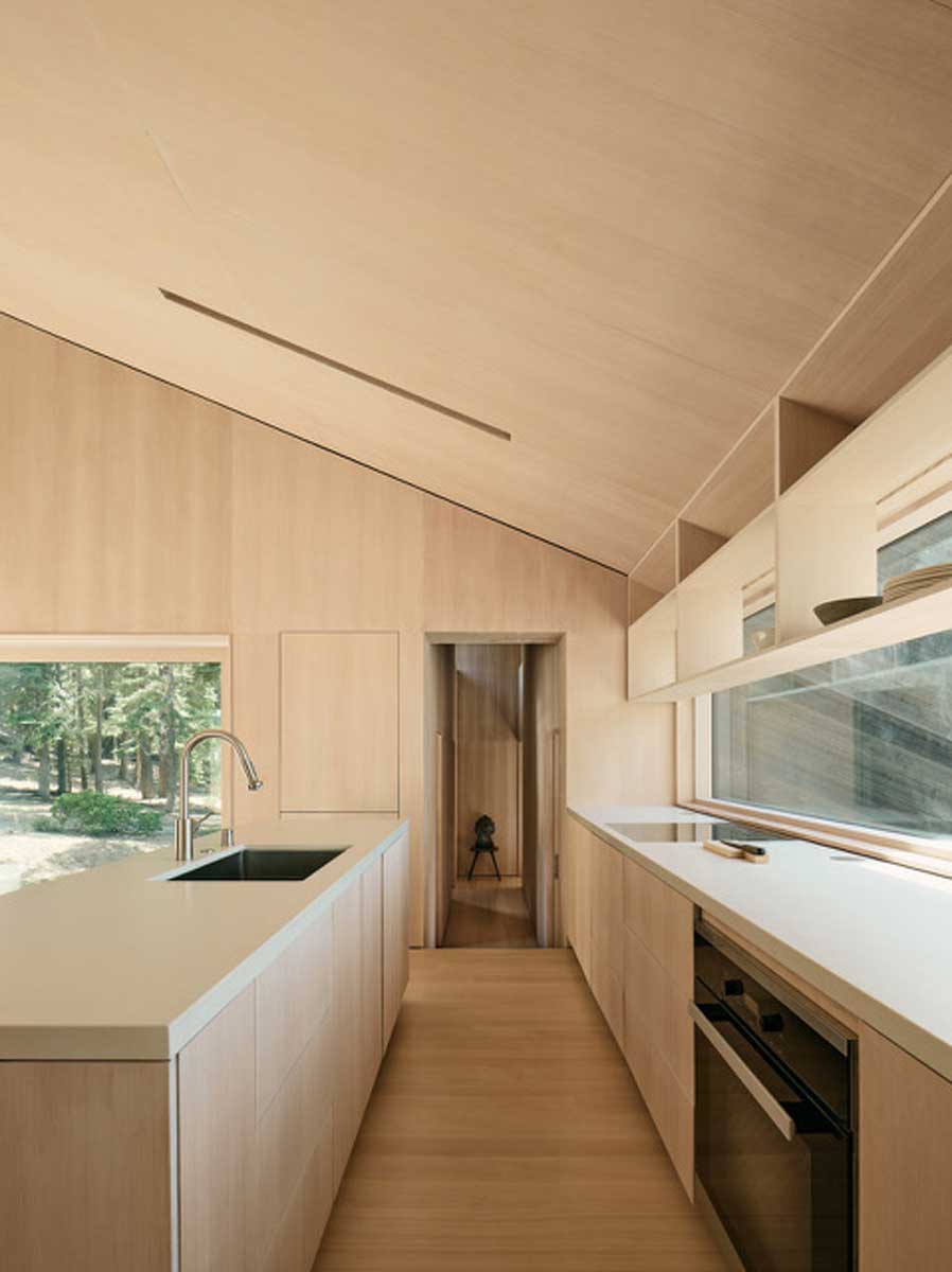 Úzká kuchyně s vestavěným nábytkem ze světlého dřeva a velkým oknem propojuje interiér s okolním lesem.
