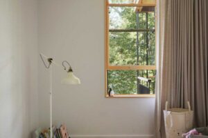 Světlá ložnice s dřevěnou podlahou, vysokým oknem s výhledem do zeleně a jemnými textiliemi v přírodních tónech.