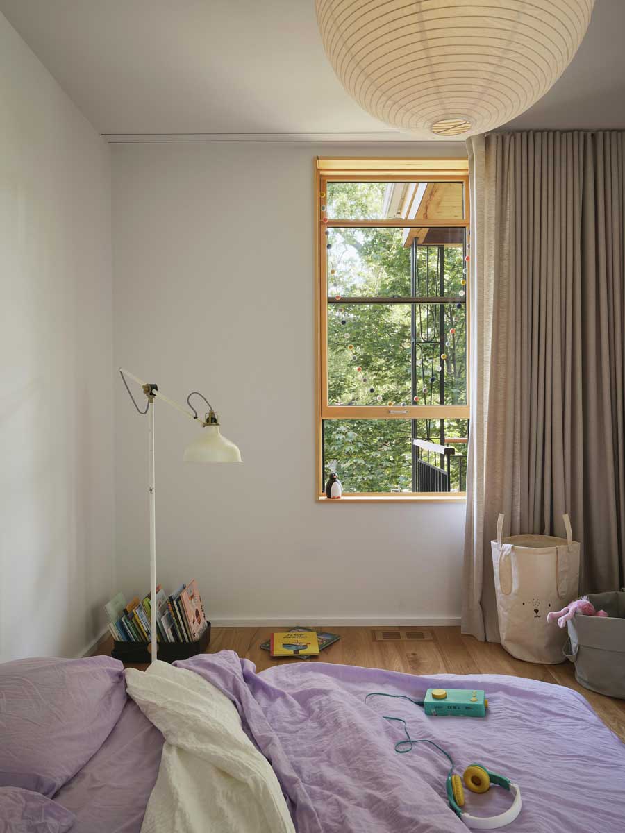 Světlá ložnice s dřevěnou podlahou, vysokým oknem s výhledem do zeleně a jemnými textiliemi v přírodních tónech.