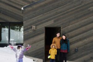 Rodina stojí před tmavou fasádou chaty zasazené do sněhu a lesního svahu.