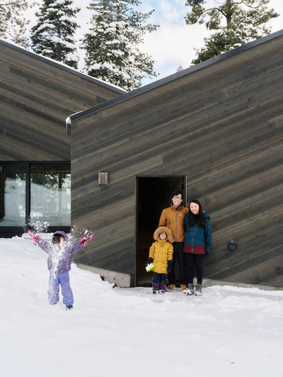 Rodina stojí před tmavou fasádou chaty zasazené do sněhu a lesního svahu.