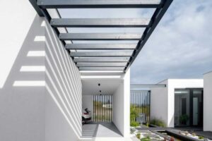 Kovová pergola s lamelovou konstrukcí vrhající stíny na světlou fasádu domu.