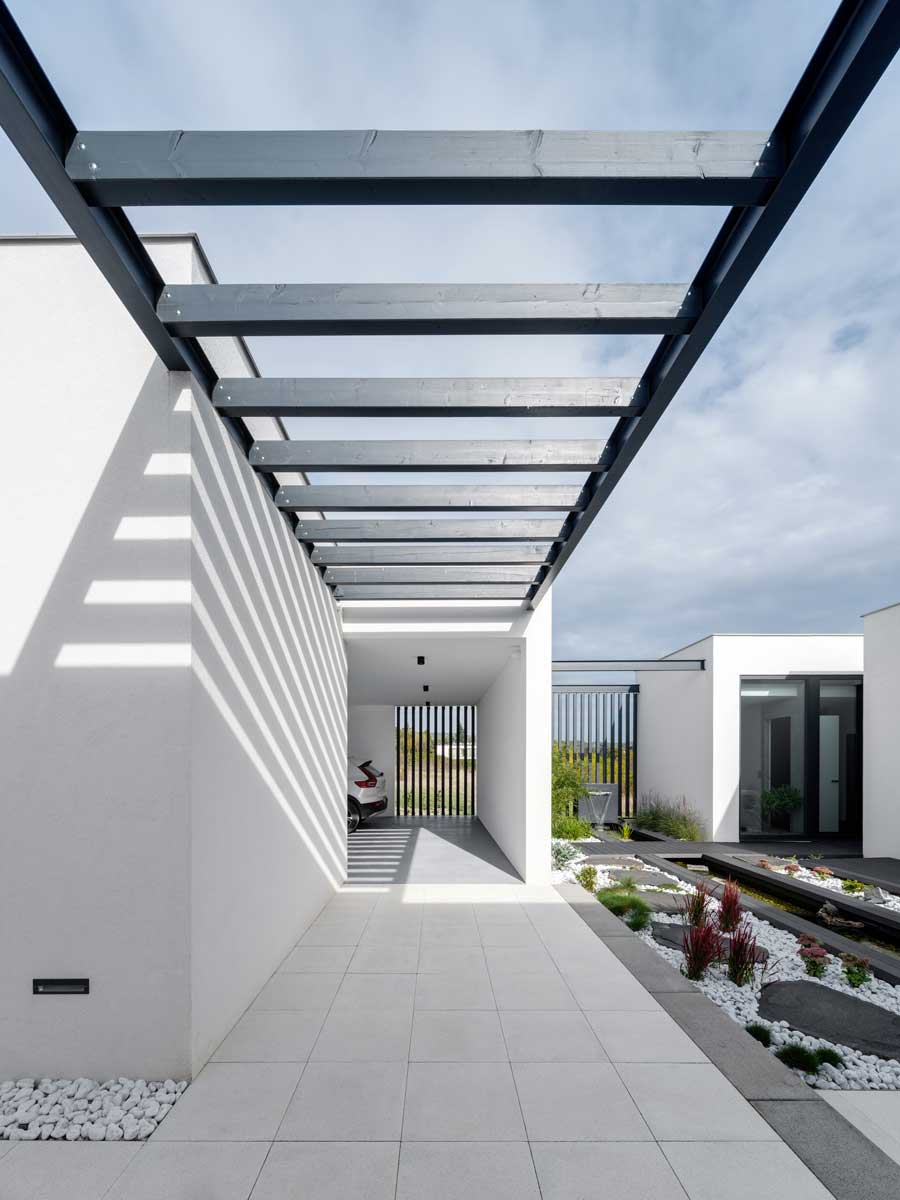 Kovová pergola s lamelovou konstrukcí vrhající stíny na světlou fasádu domu.