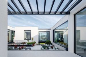 Moderní dům s plochou střechou, krytou pergolou a minimalistickou zahradou s kamennými prvky a bílým štěrkem.