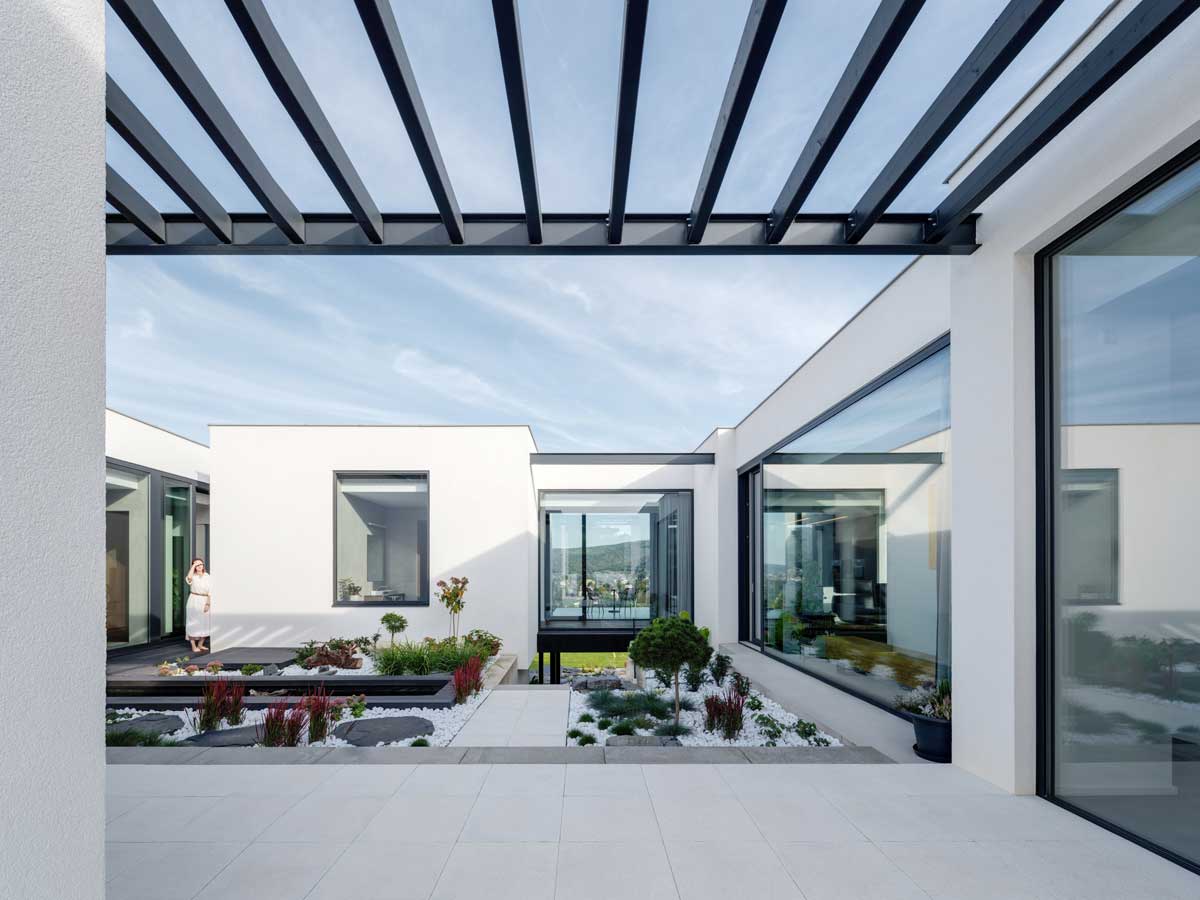 Moderní dům s plochou střechou, krytou pergolou a minimalistickou zahradou s kamennými prvky a bílým štěrkem.