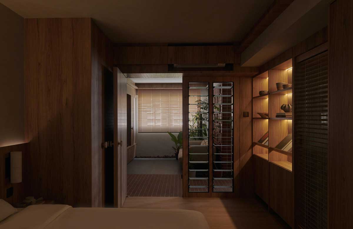 Pohled z ložnice přes dřevěné rámy a lamely odhaluje cihlové prvky a zeleň v centrální obytné části bytu.