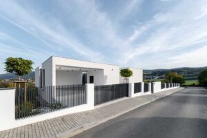 Bílý moderní dům s jednoduchou fasádou a kovovým plotem při pohledu z ulice.