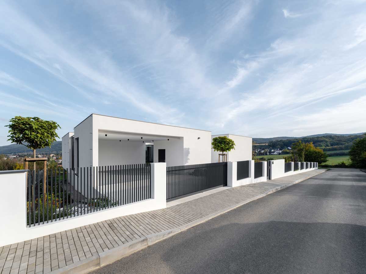Bílý moderní dům s jednoduchou fasádou a kovovým plotem při pohledu z ulice.