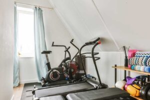 Podkrovní cvičební kout s běžeckým pásem, rotopedem a posilovací lavicí využívá přirozené světlo ze střešního okna a kompaktní uspořádání pod šikminou.