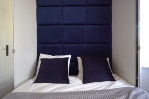Postel v malé ložnici s čalouněným tmavě modrým čelem a úsporným řešením prostoru