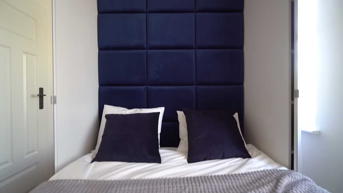 Postel v malé ložnici s čalouněným tmavě modrým čelem a úsporným řešením prostoru