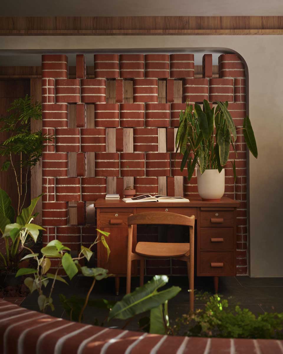 Pracovní kout s dřevěným psacím stolem stojí před dekorativní stěnou z obloukových cihel a je obklopen pokojovými rostlinami.