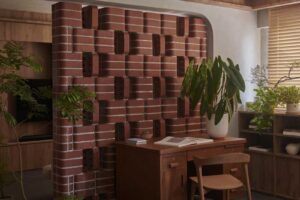 Dřevěný pracovní stůl s jednoduchou židlí je zasazen mezi zeleň a výraznou cihlovou stěnu s plastickým vzorem.