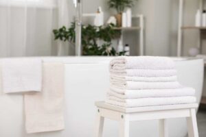 Komínek bílých ručníků položený na stoličce v moderní koupelně s vanou v pozadí.