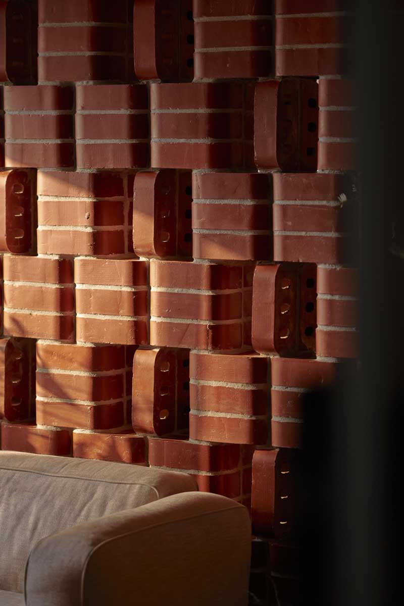 Detail plastické cihlové stěny v kombinaci s čalouněnou sedačkou zdůrazňuje strukturu materiálu a hru světla.