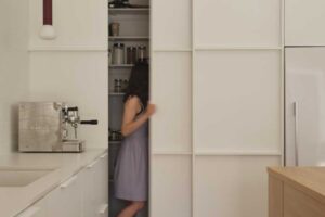 Kuchyň s posuvnými dveřmi do skryté spíže a vestavěnými bílými skříněmi.