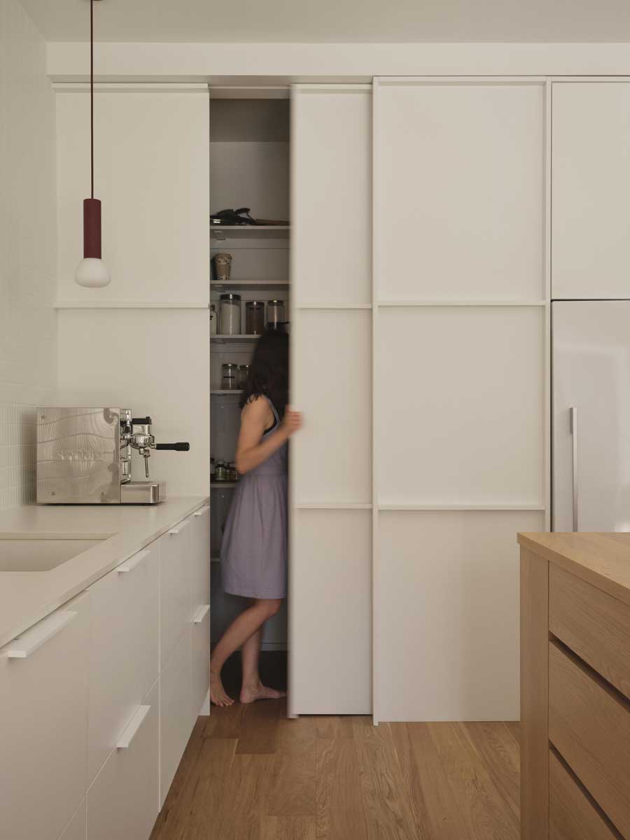 Kuchyň s posuvnými dveřmi do skryté spíže a vestavěnými bílými skříněmi.