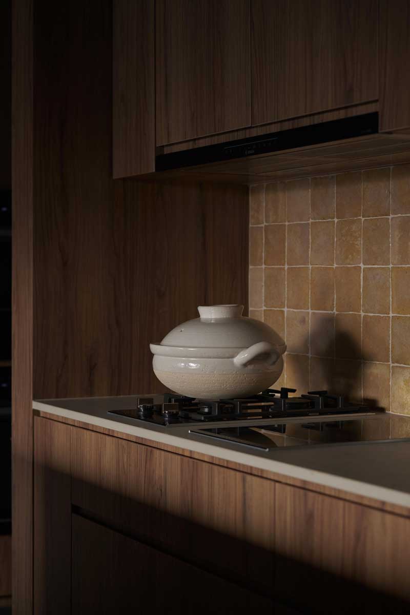 Kuchyňský sporák s keramickým hrncem je zasazen do dřevěné linky s obkladem z malých čtvercových dlaždic v teplém odstínu.