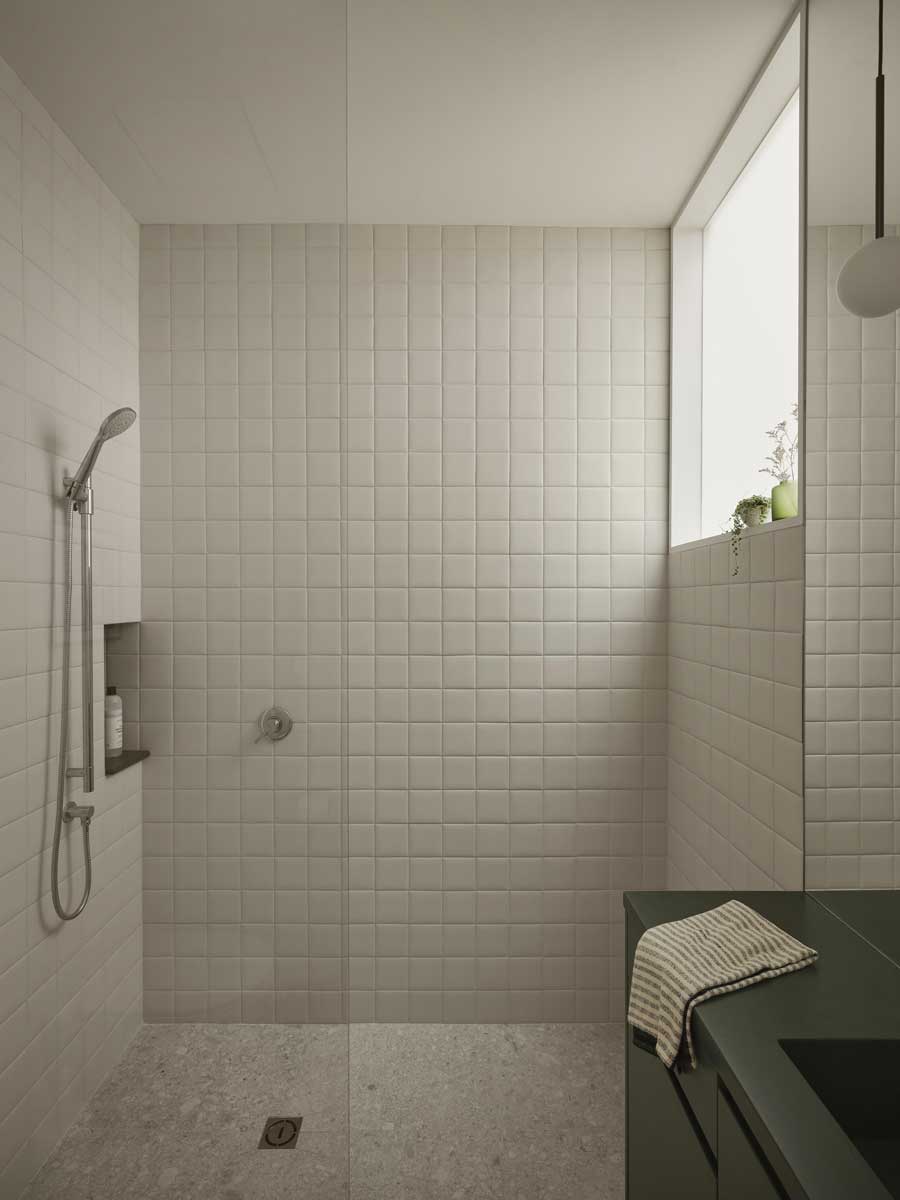 Minimalistický sprchový kout s bílými obklady, nikou na kosmetiku a velkým oknem pro přirozené světlo.