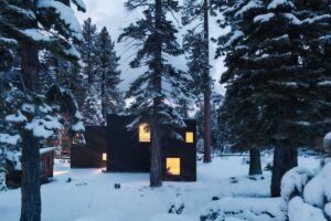 Chata s osvětlenými okny působí v zimním lese jako klidné a teplé útočiště.