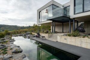 Rodinný dům s proskleným balkonem a přírodním koupacím jezírkem v zahradě.