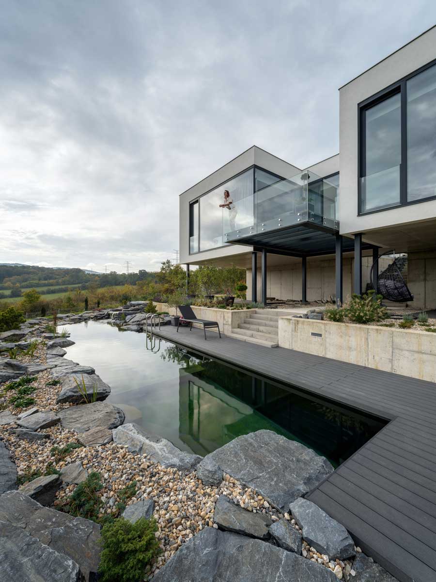 Rodinný dům s proskleným balkonem a přírodním koupacím jezírkem v zahradě.