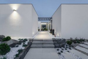 Minimalistický vstupní chodník mezi dvěma bílými hmotami domu s kamennými zídkami a bílým kačírkem.