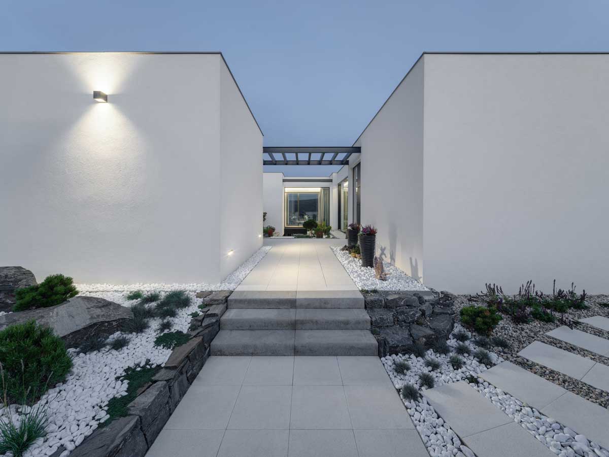 Minimalistický vstupní chodník mezi dvěma bílými hmotami domu s kamennými zídkami a bílým kačírkem.