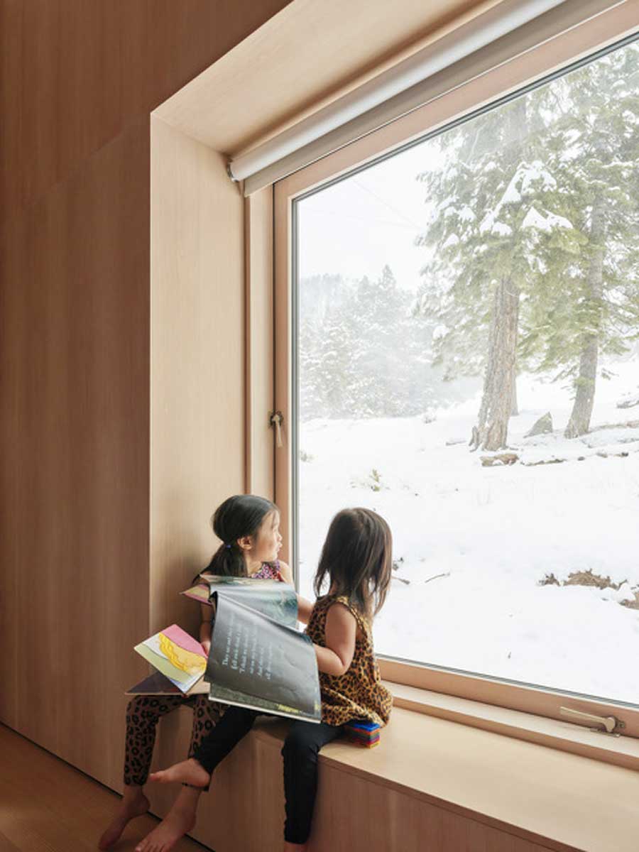 ěti sedí na vestavěné lavici u velkého okna a sledují zasněžený les za sklem.