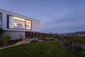 Moderní vila na svahu při večerním světle s osvětleným interiérem a upravenou zahradou.