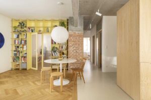 Otevřený obytný prostor s výraznou žlutou knihovnou, jídelním stolem a přiznanou cihlovou stěnou po rekonstrukci bytu