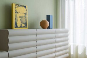 Designová komoda se zaobleným rytmickým profilem stojí u okna a doplňuje ji barevné dekorace v modré, žluté a terakotové.