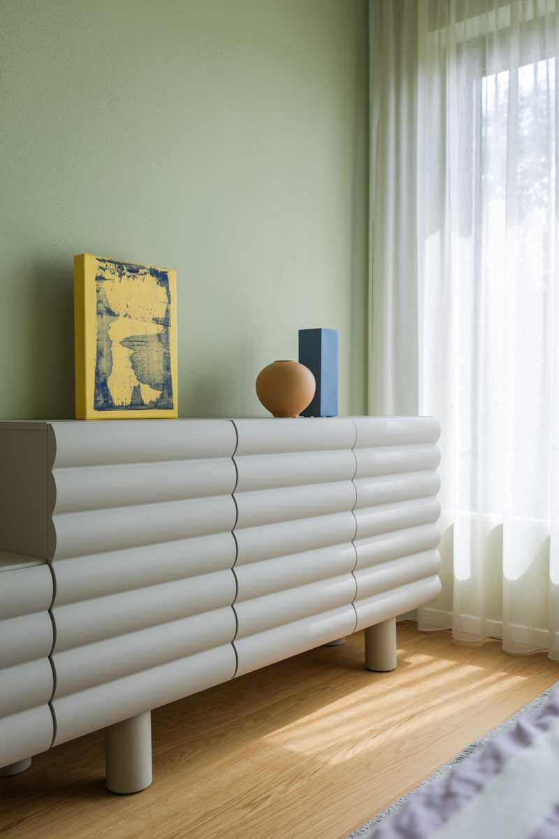 Designová komoda se zaobleným rytmickým profilem stojí u okna a doplňuje ji barevné dekorace v modré, žluté a terakotové.