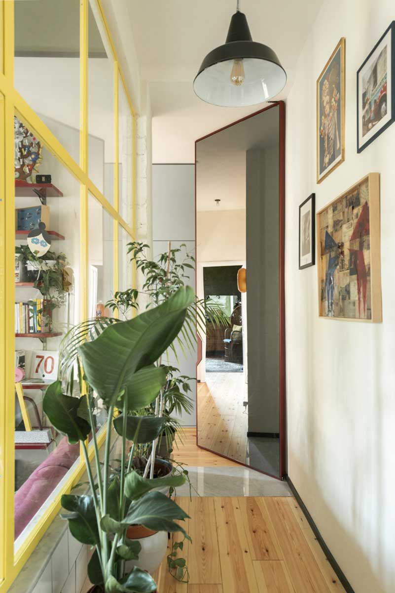 Úzká chodba s rostlinami velkým zrcadlem a prosklenou příčkou se žlutými rámy.