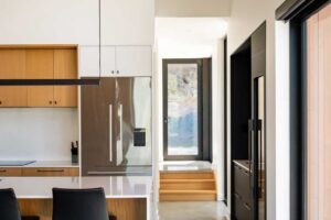 Minimalistická kuchyň s dřevěnými prvky a vysokým stropem propojená s chodbou a vstupem.