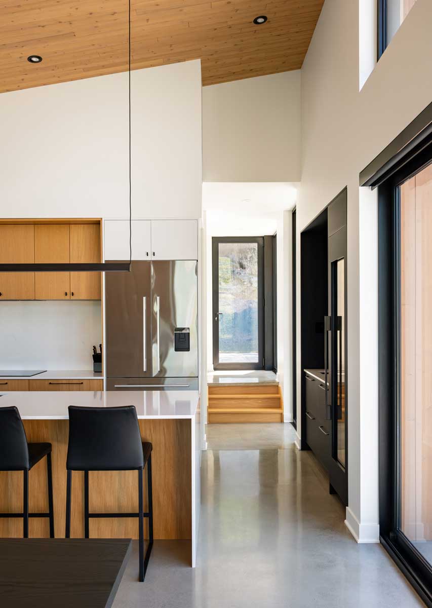 Minimalistická kuchyň s dřevěnými prvky a vysokým stropem propojená s chodbou a vstupem.